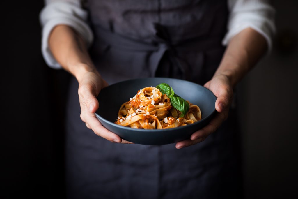 aubergine and sun dried tomato tagliatelle pasta in blue dish held by women in blue apron.