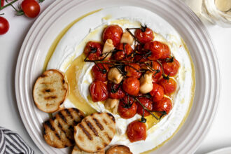 Confit tomatoes on whipped cheese with grilled baguette on a plate surrounded by cups of wine and fresh tomatoes.